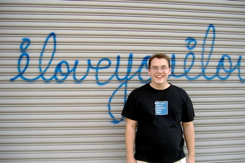 Photograph of a young, smiling, caucuasian man standing in front of a closed garage door. Spray-painted on the door, in blue, is grafitti reading 'I love you'.